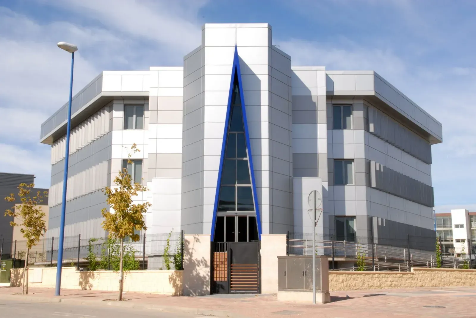 Un moderno edificio de oficinas de tres plantas con una entrada central triangular de cristal con detalles azules y revestimiento exterior de metal gris.