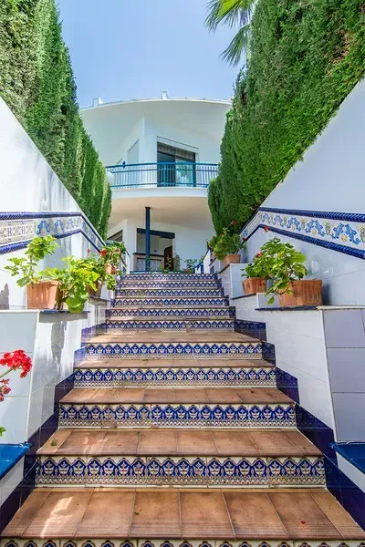 Una escalera de piedra con contrahuellas de azulejos decorativos azules y blancos conduce a una casa blanca enmarcada por altos setos verdes.