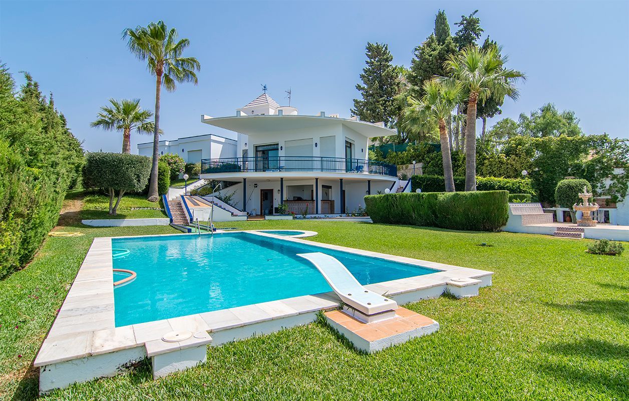 Una villa de un blanco brillante con una piscina de color turquesa, un trampolín y palmeras, rodeada de un exuberante césped verde.
