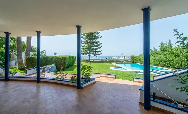Un patio cubierto con suelo de baldosas con vistas a un jardín trasero con césped, piscina y vistas lejanas al océano.