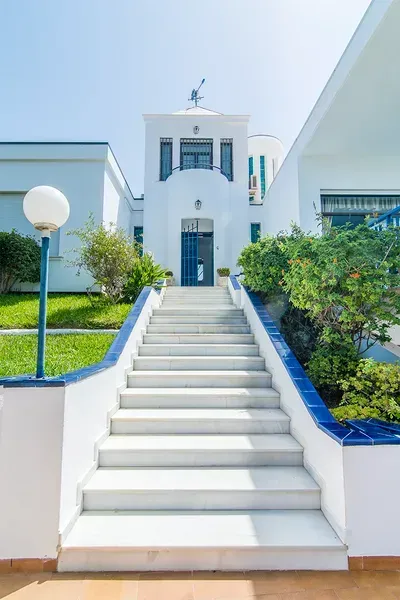 Una amplia escalera blanca con detalles de azulejos azules conduce a la entrada de un edificio blanco bajo un cielo azul despejado.