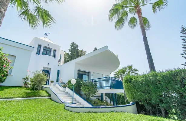 Una casa blanca con un moderno tejado curvo y detalles azules, rodeada de un césped verde y palmeras bajo un cielo azul brillante.