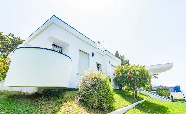 Vista desde un ángulo bajo de una casa blanca y moderna con un balcón curvo, rodeada de césped verde y un arbusto florido de colores vivos.