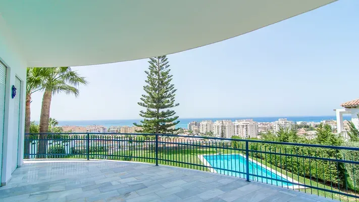 Un amplio balcón con suelo de baldosas con vistas a un césped verde, una piscina, edificios a lo lejos y el mar.