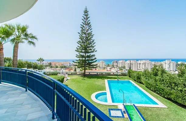 Desde un balcón se puede contemplar una piscina de color azul brillante en un jardín con césped, palmeras, edificios costeros y el mar.