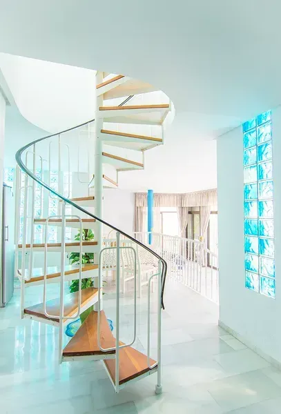 Una escalera de caracol con barandillas blancas y escalones de madera se alza en una habitación blanca y luminosa, junto a una pared de bloques de vidrio azul.