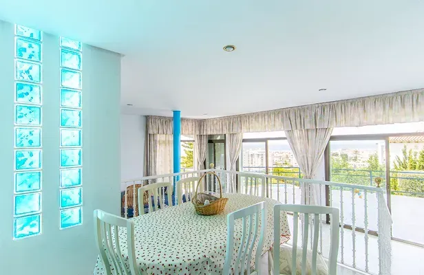 Comedor con mesa, sillas blancas, pared de bloques de vidrio y grandes ventanales con cortinas color crema con vistas al exterior.