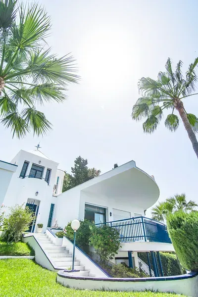 Una villa blanca de estilo mediterráneo con detalles azules, que cuenta con balcón y escaleras, rodeada de palmeras y vegetación.