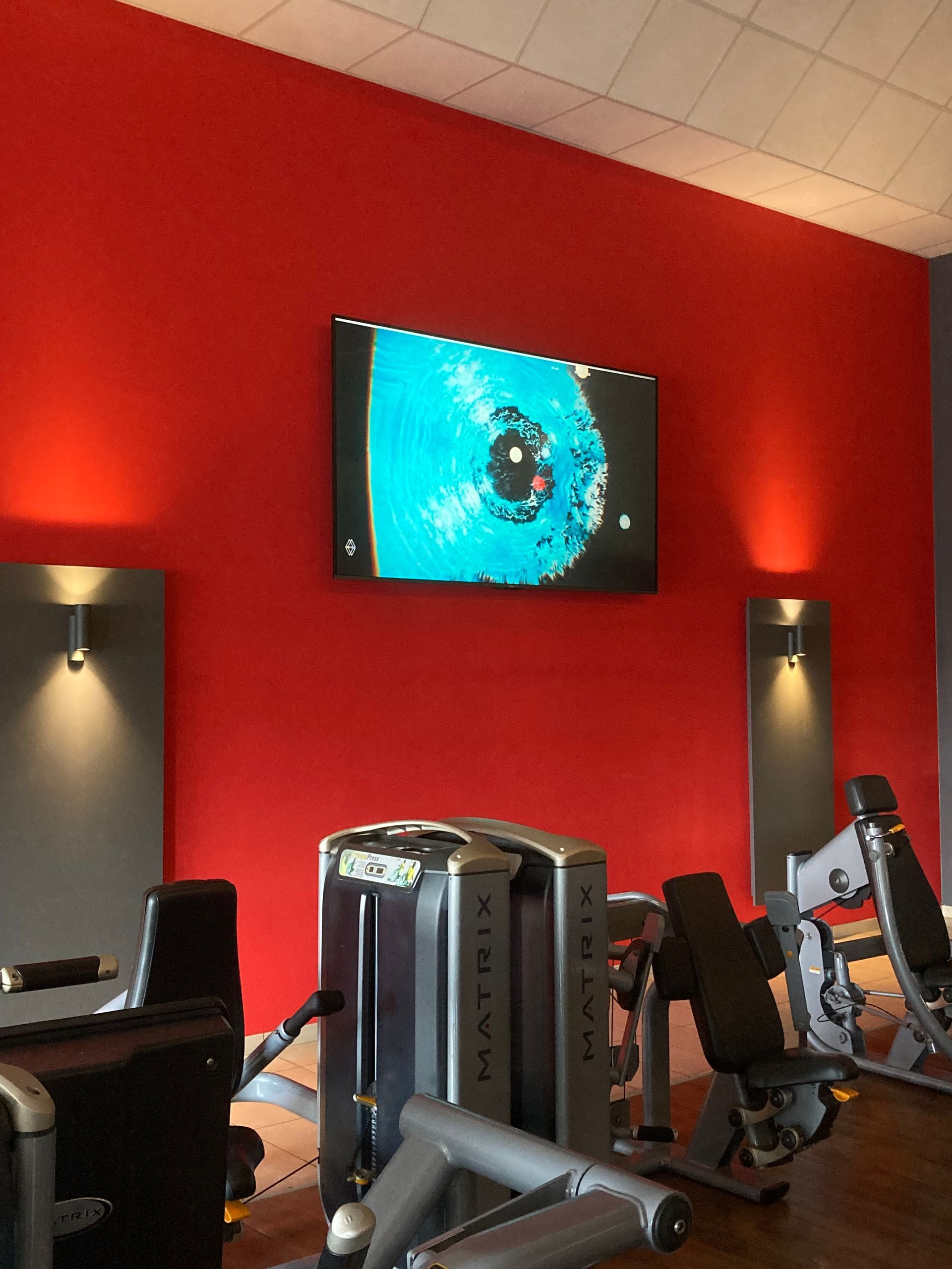 Intérieur de salle de sport avec mur rouge, télévision, équipement d'exercice et éclairage mural.