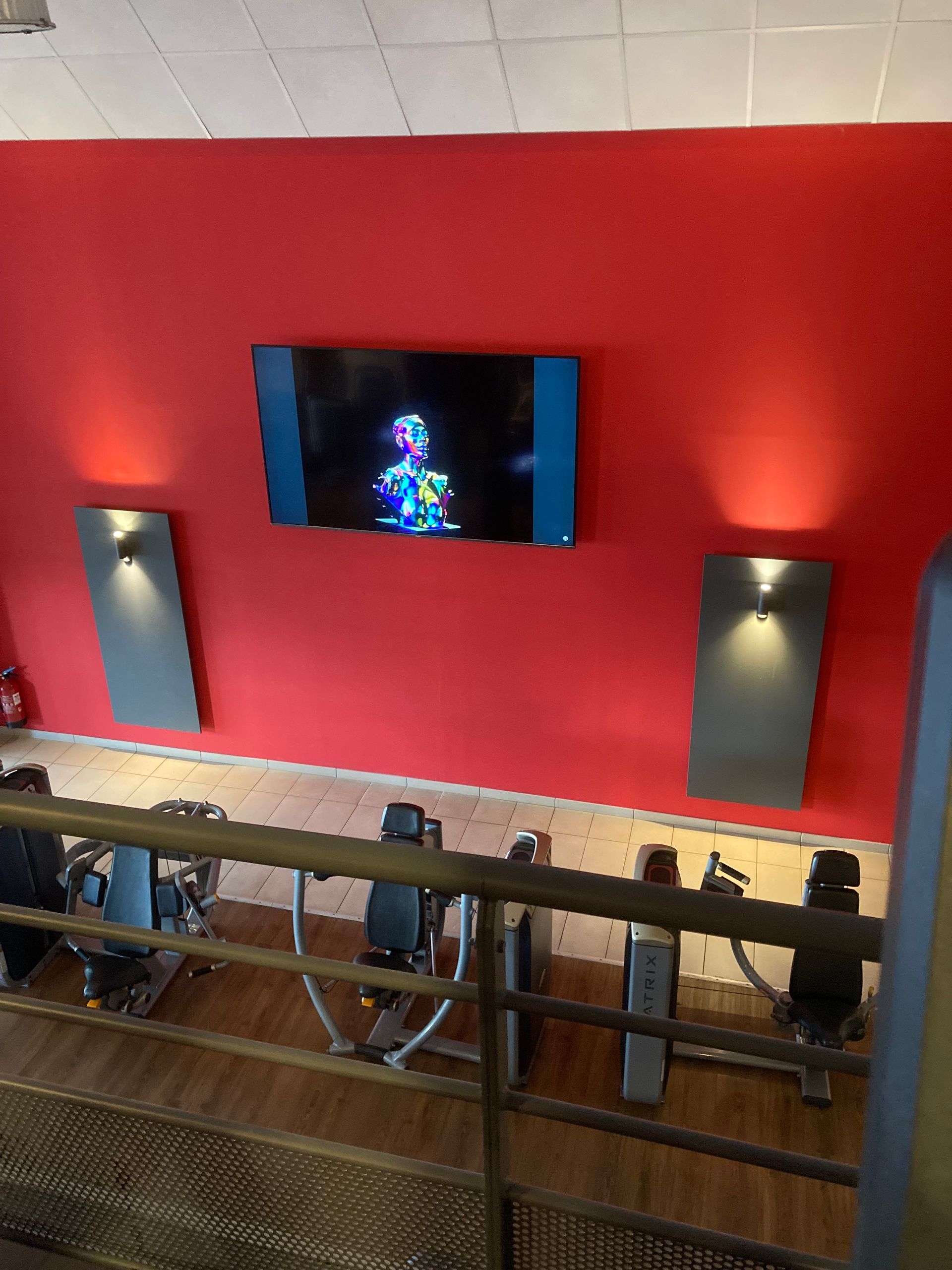 Intérieur de la salle de sport : mur rouge avec télévision et deux appliques. Appareils d'exercice visibles en dessous.