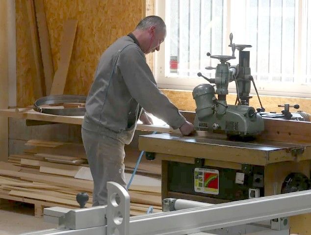 Un homme en tenue grise travaille sur une machine dans un atelier où du bois est empilé et où la lumière du soleil entre par une fenêtre.