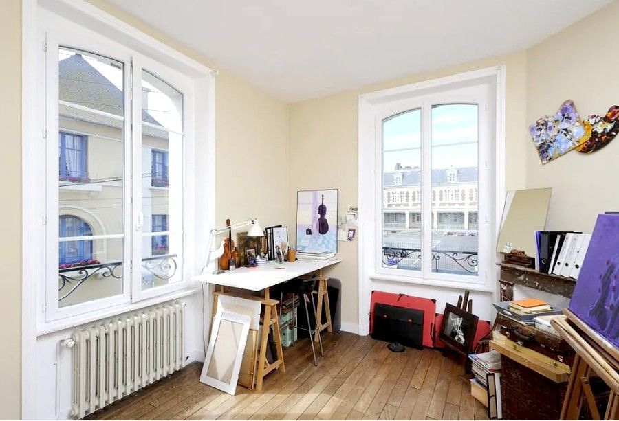 Chambre d'angle lumineuse avec de grandes fenêtres, parquet et un bureau avec du matériel d'art et des instruments