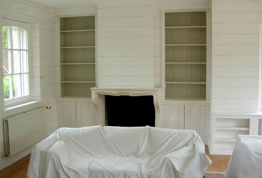 Salon d'un blanc éclatant avec étagères intégrées, cheminée et meubles recouverts de draps blancs