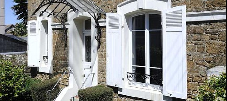 Volets blancs sur une maison en pierre, fenêtres ouvertes et petit auvent au-dessus de la porte