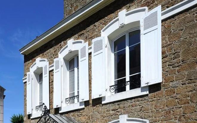 Façade en pierre d'une maison avec volets blancs et balcon se détachant sur un ciel bleu.