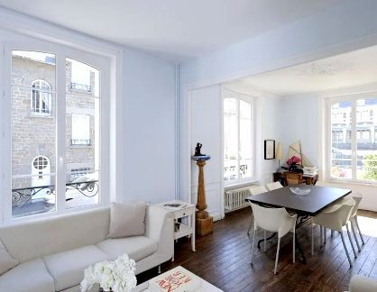 Salon-salle à manger lumineux et aéré avec canapé blanc, parquet foncé et table près de grandes fenêtres.