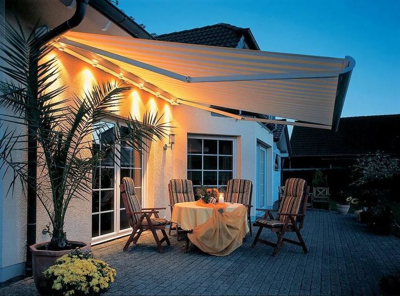 Patio au crépuscule avec auvent éclairé, table dressée pour le dîner, chaises et palmier en pot près d'une maison