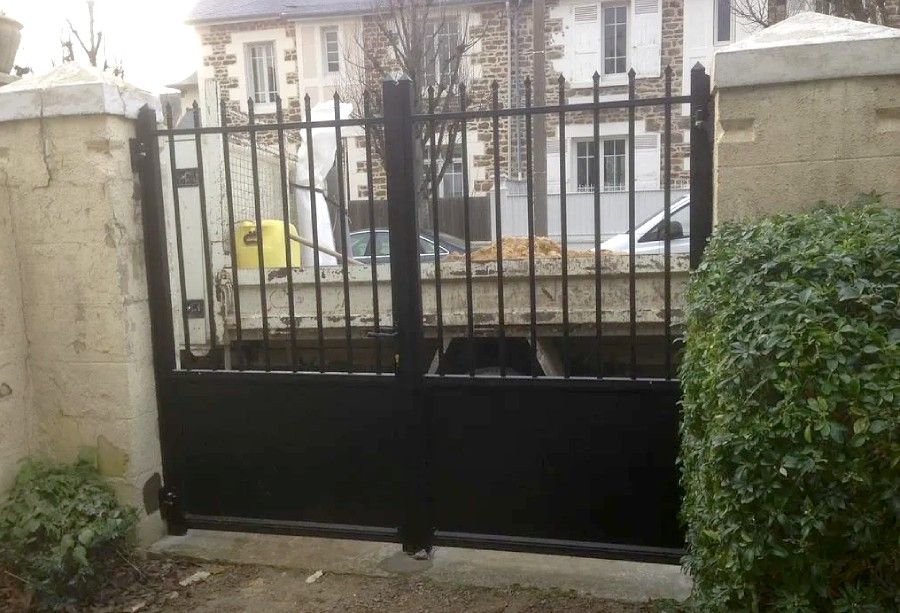 Portail en métal noir devant l'entrée d'une maison entourée de murs en pierre et d'arbustes.