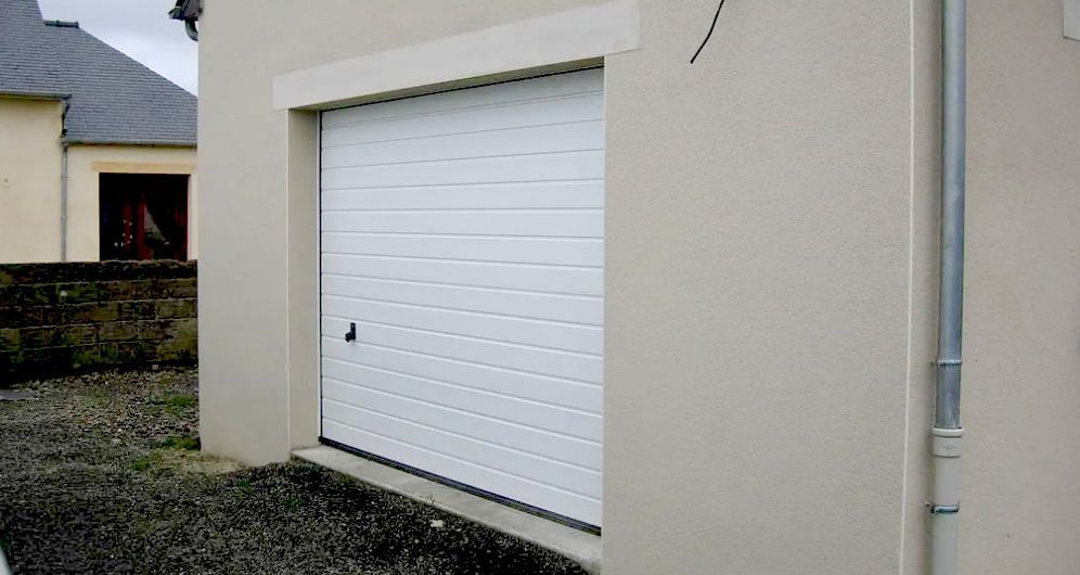 Porte de garage blanche sur une façade de maison beige, à côté d'une allée de gravier.