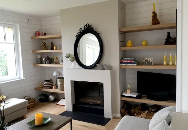 Salon lumineux avec cheminée, étagères intégrées, miroir et banquette beige près d'une fenêtre.