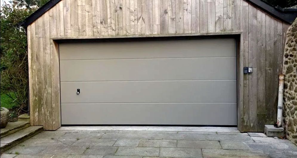 Porte de garage grise fermée sur un bâtiment en bois patiné par le temps, avec une allée en pierre.