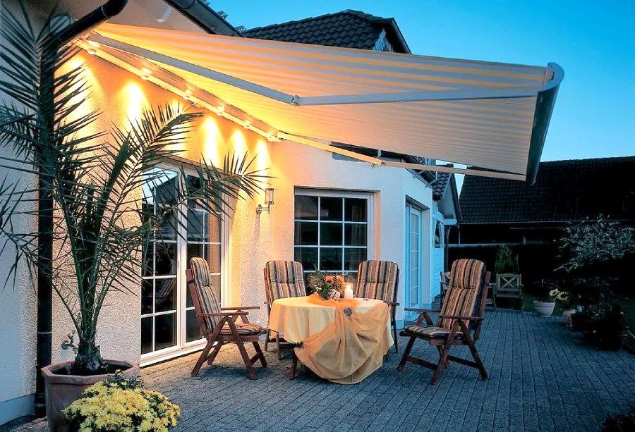 Terrasse aménagée sous une pergola blanche illuminée d'une douce lumière, avec une table ronde et des chaises en bois au crépuscule