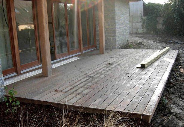 Terrasse en bois jouxtant une maison aux grandes portes vitrées et au mur de pierre, bordée de terre et de buissons.