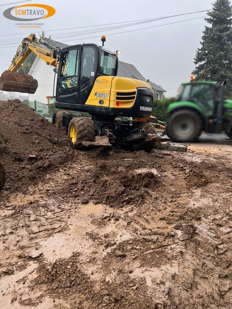 Une excavatrice jaune déplace de la terre dans un champ boueux à côté d'un tracteur vert.
