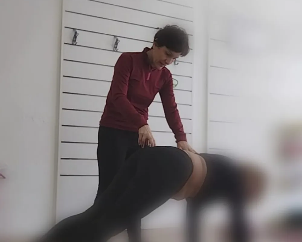 Una persona ayuda a otra a realizar un ejercicio de estiramiento en el interior, junto a una pared con líneas horizontales.