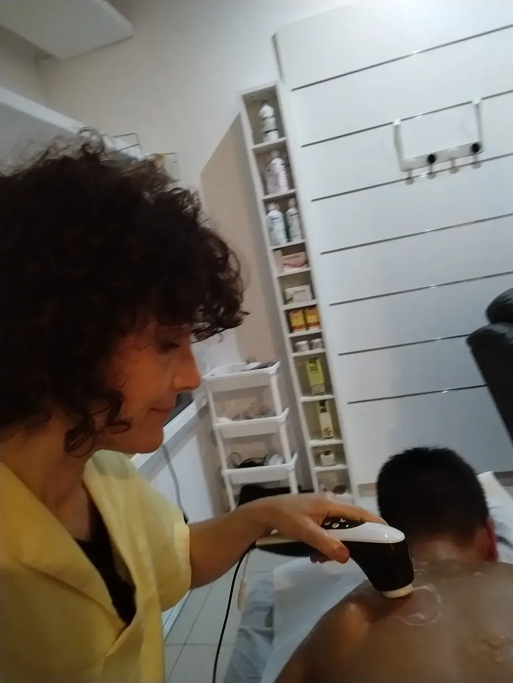 Una persona con una blusa amarilla usa un dispositivo portátil en la espalda de otra persona en una habitación luminosa.
