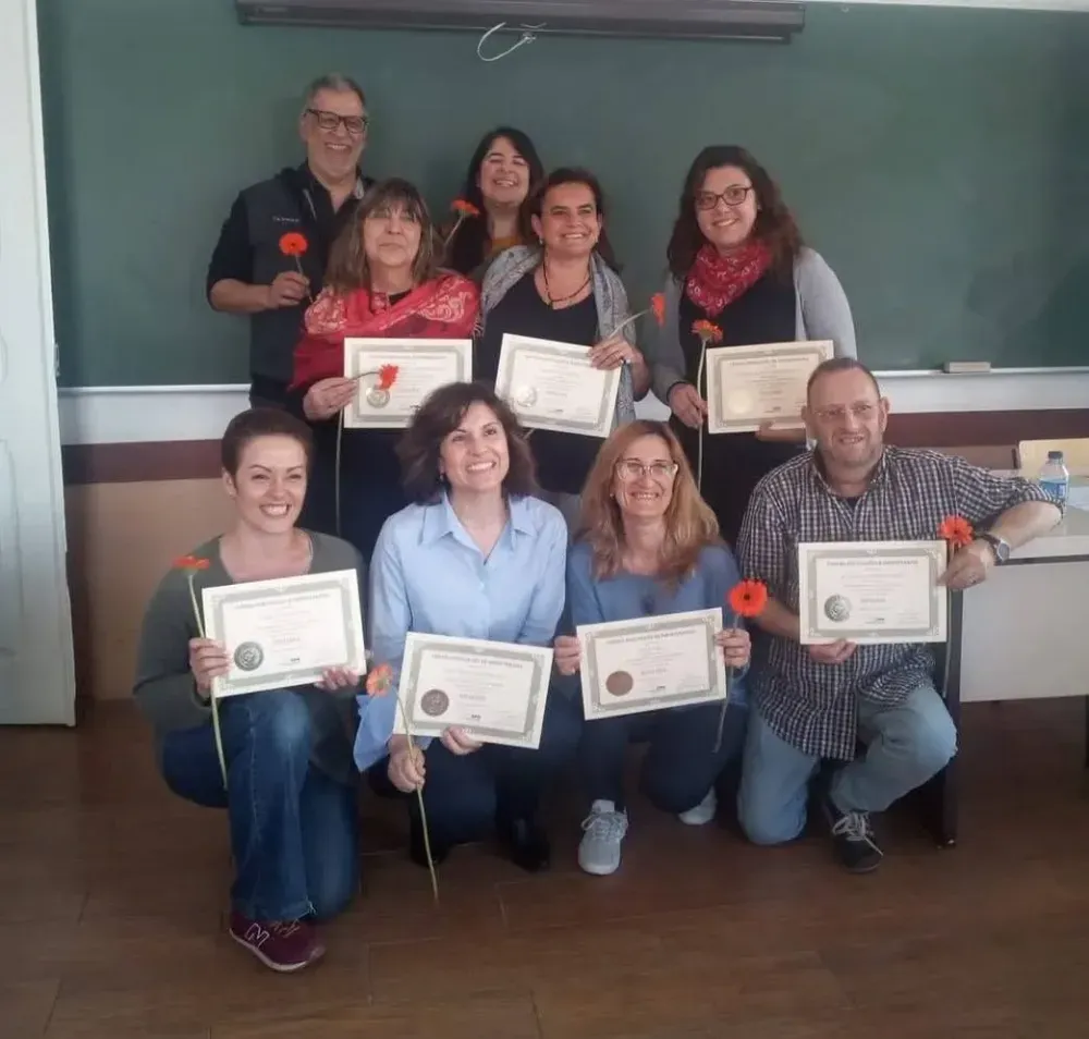 Grupo de personas sosteniendo certificados y flores, posando en un aula con una pizarra de fondo.