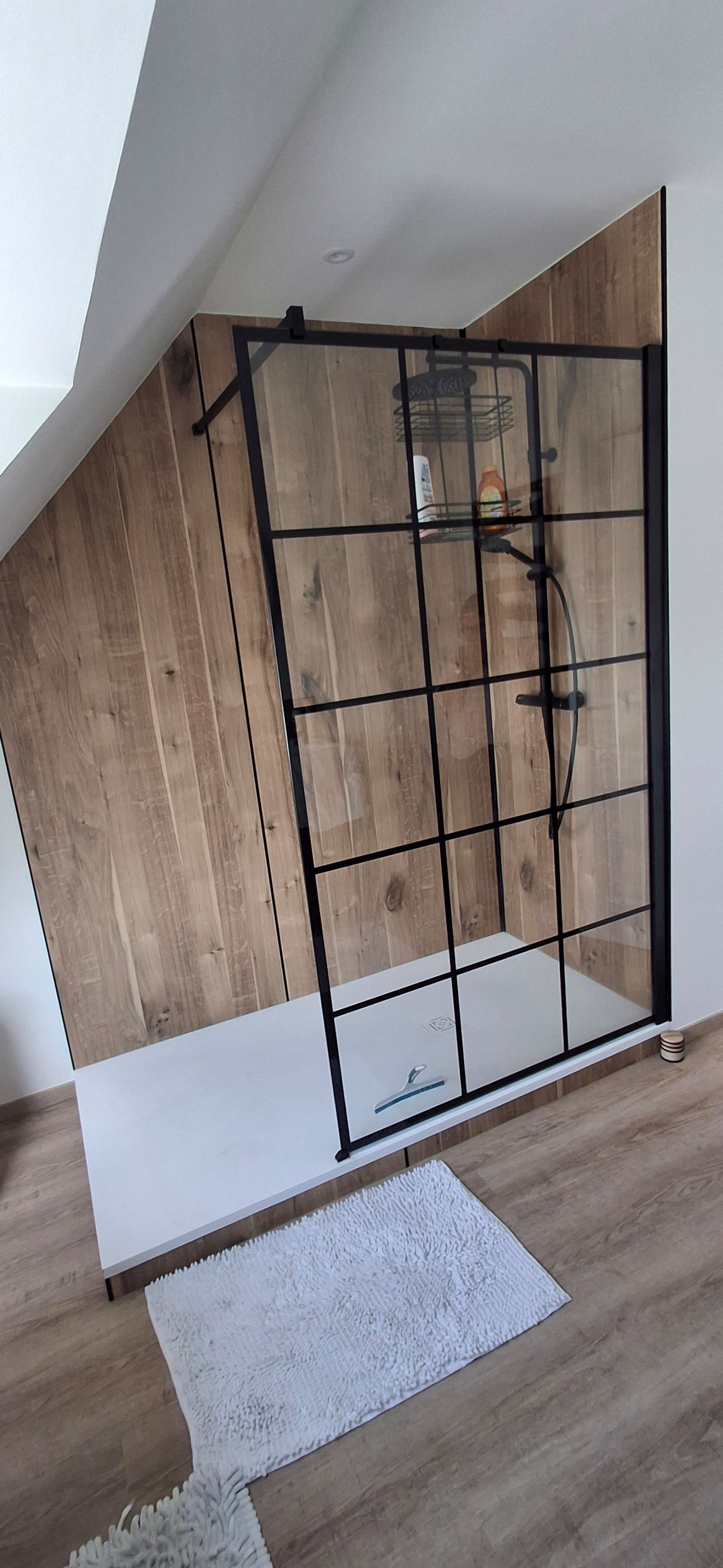 Cabine de douche avec grille noire, mur lambrissé et tapis blanc.