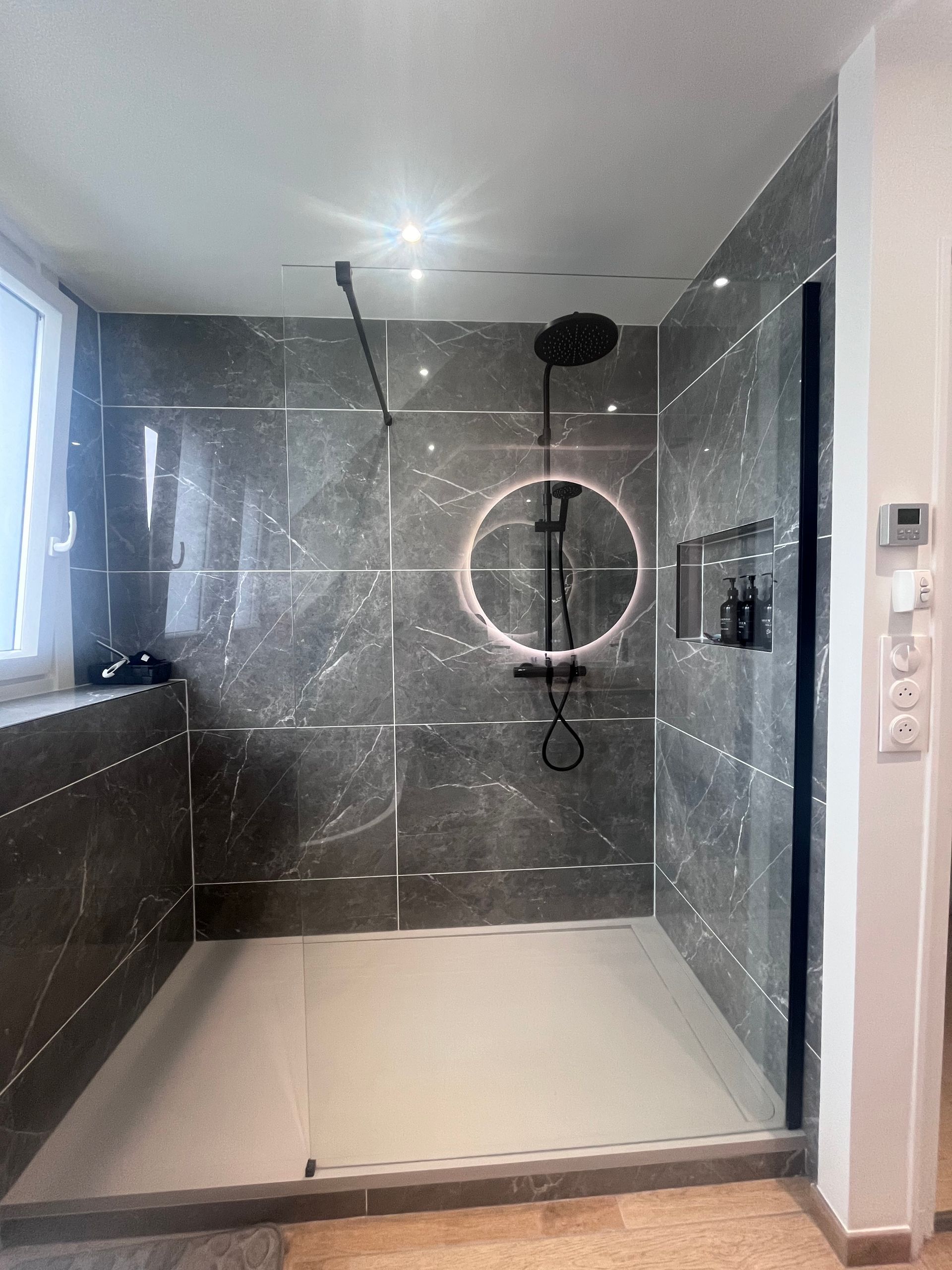 Douche carrelée grise avec des accessoires noirs et un miroir lumineux.