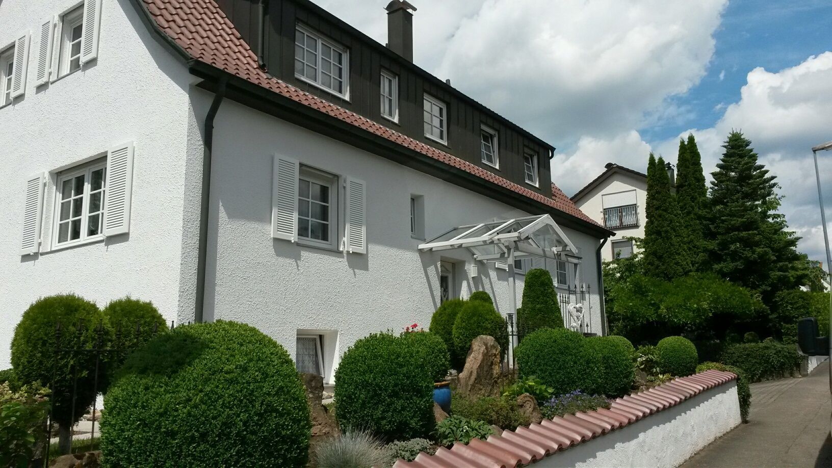 Ein weißes Haus mit einem roten Ziegeldach ist von Büschen und Bäumen umgeben.