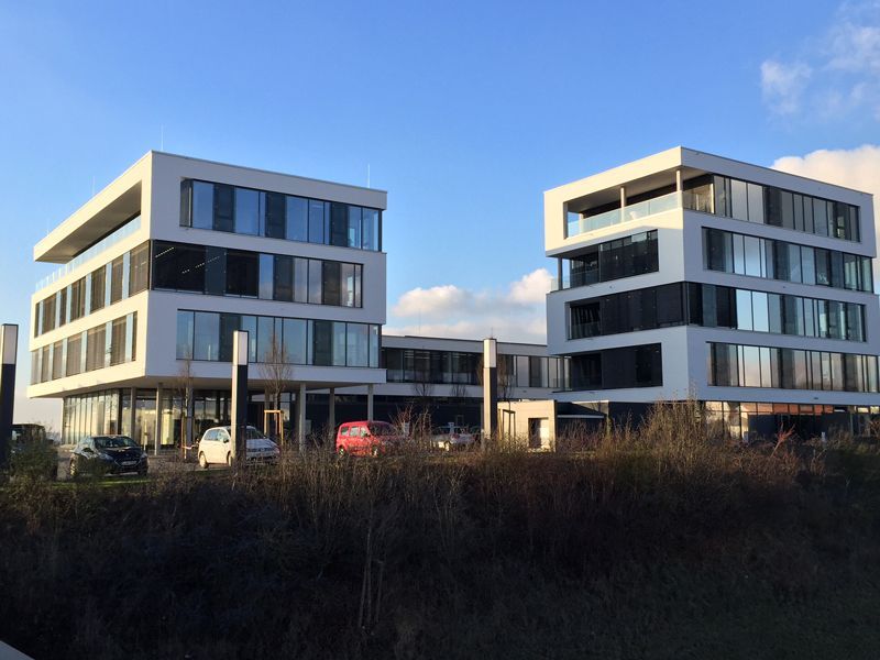 Auf einem Feld stehen zwei Gebäude nebeneinander. Davor sind Autos geparkt.