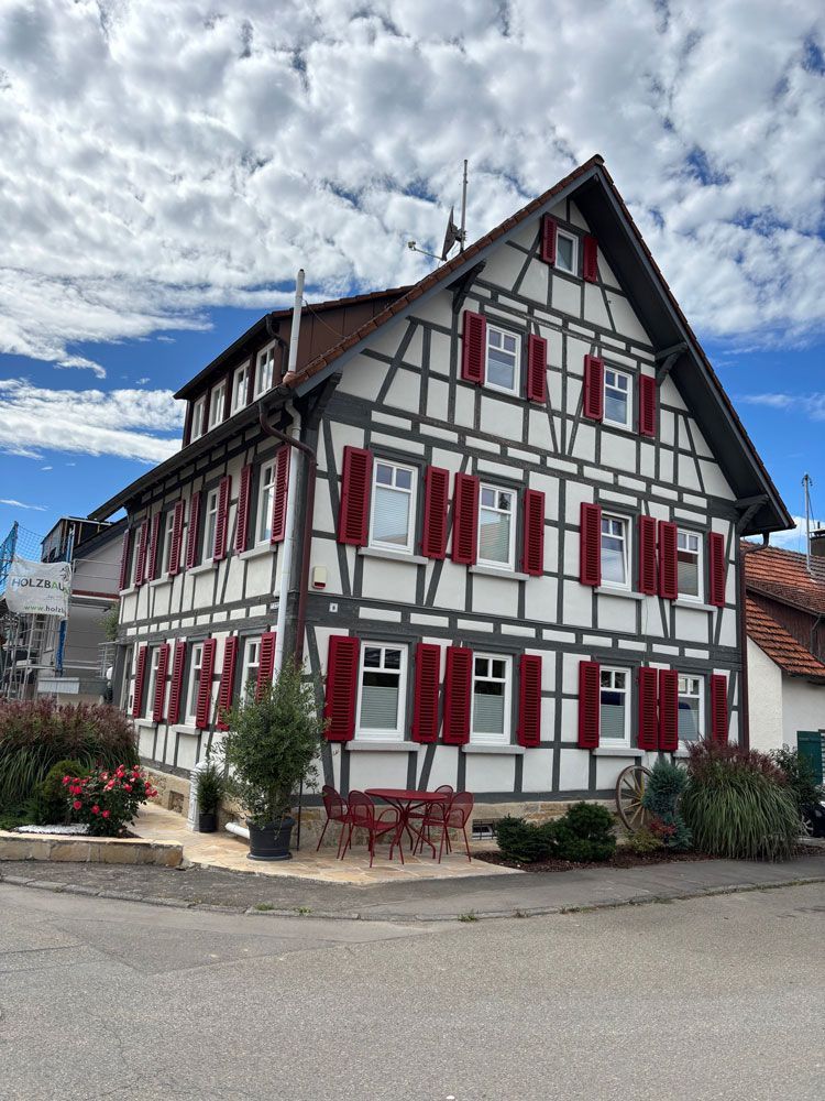 Fachwerkhaus mit roten Fensterläden auf einem Eckgrundstück unter bewölktem Himmel.