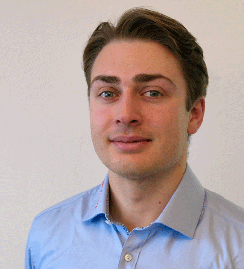 Ein Mann in einem blauen Hemd steht vor einer Tür und schaut in die Kamera.