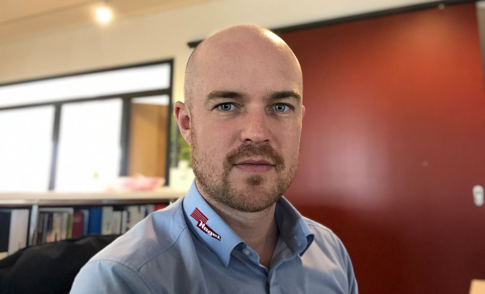 Ein Mann in einem blauen Hemd steht vor einer Tür und schaut in die Kamera.