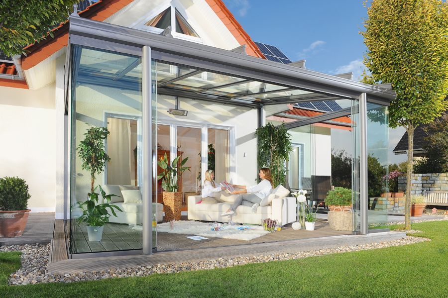 Eine Frau sitzt auf einem Sofa unter einer glasüberdachten Terrasse vor einem Haus.