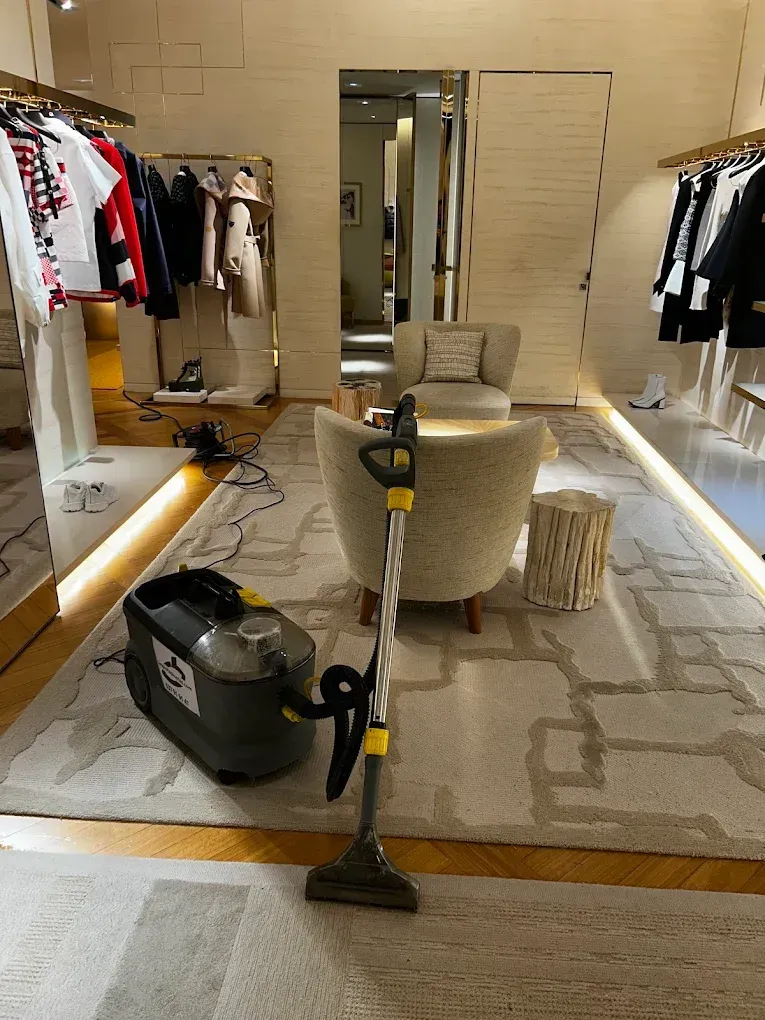Una alfombra siendo limpiada en una tienda de ropa, con una aspiradora y un sillón beige a la vista.