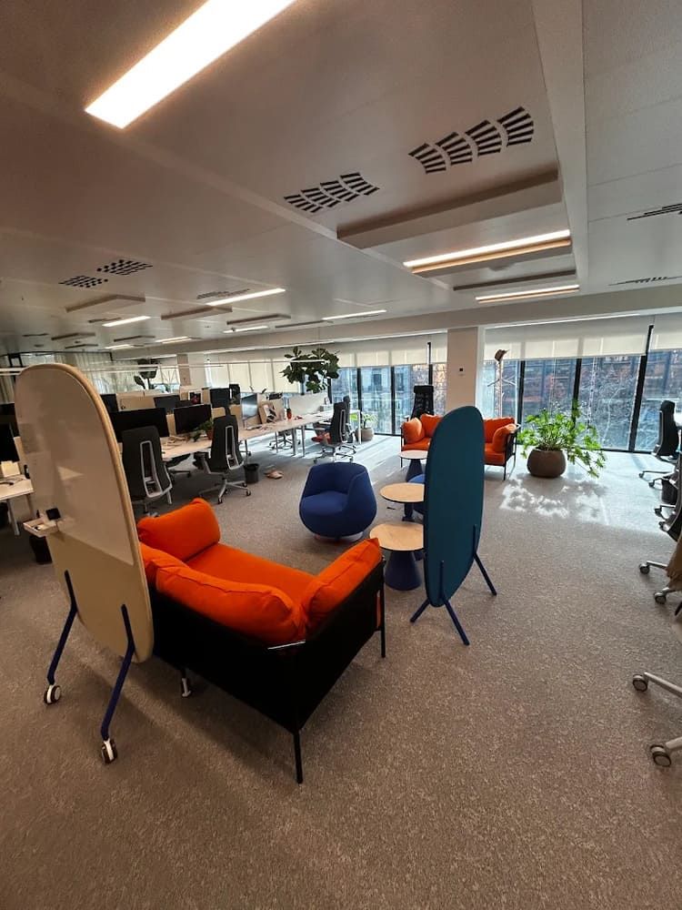 Oficina diáfana con sofá naranja, sillas azules y mesas redondas, bajo una iluminación brillante. Ventanas y escritorios al fondo.