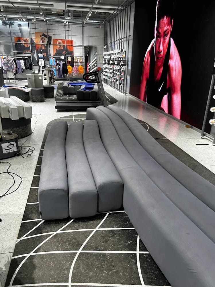 Dentro de una tienda, un banco gris curvo reposa sobre un suelo de baldosas blancas y negras. Al fondo, se ve una gran representación de una persona.