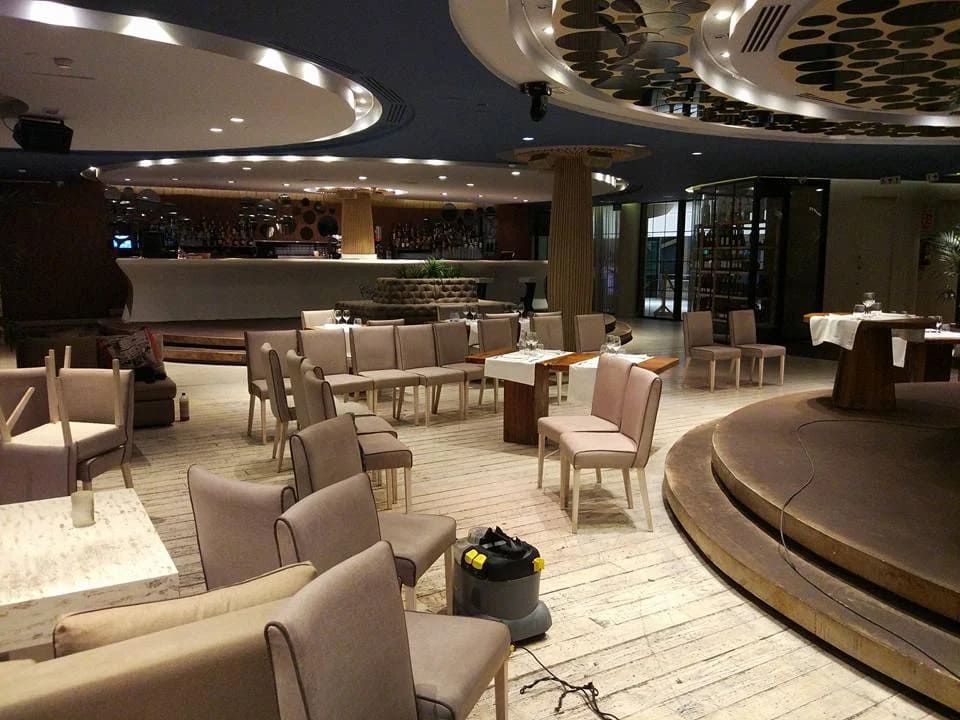 Espacio vacío para eventos con sillas dispuestas en filas alrededor de un pequeño escenario. Suelo de madera, colores neutros.