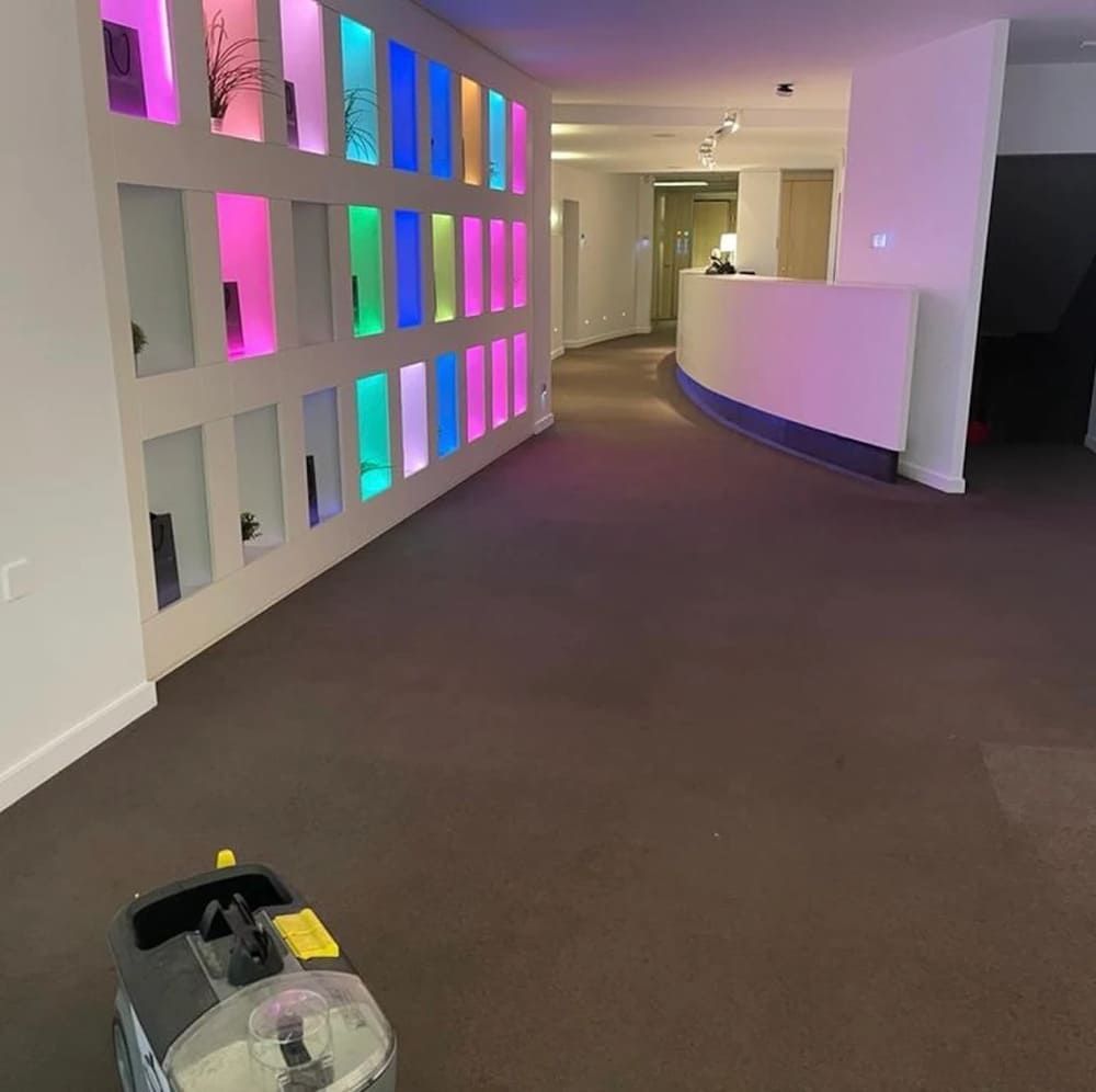 Un área de recepción de oficina alfombrada con una pared iluminada multicolor y una máquina de limpieza.