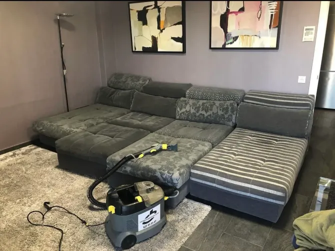 Una máquina profesional de limpieza de tapicerías reposa sobre una alfombra frente a un gran sofá modular seccional gris.