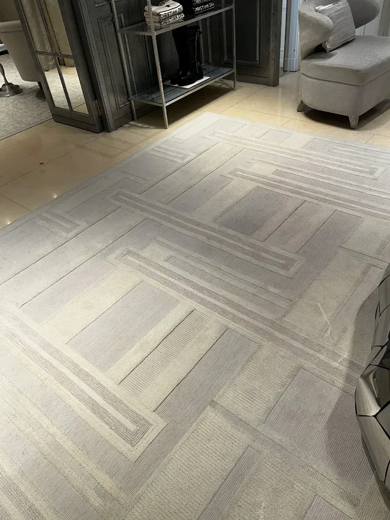 Una alfombra gris y crema con dibujos geométricos sobre un suelo pulido; se ven una estantería plateada y una otomana tapizada.