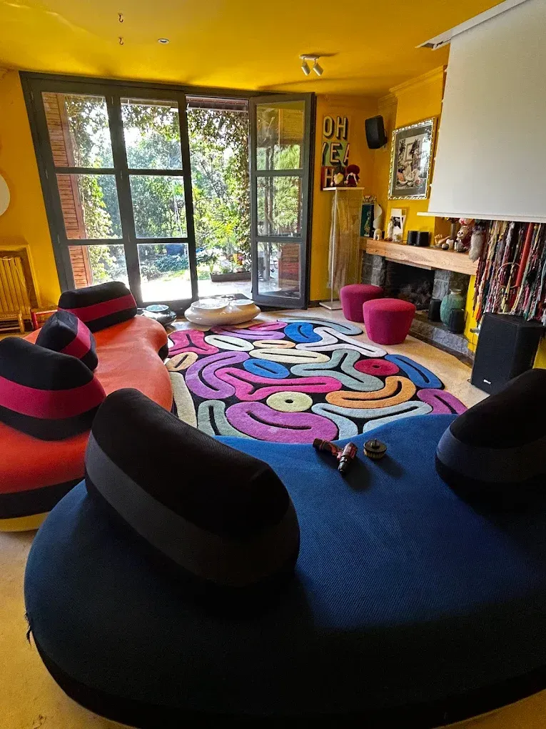 Sala de estar colorida con muebles únicos, alfombra estampada y grandes ventanales con vistas a los árboles.