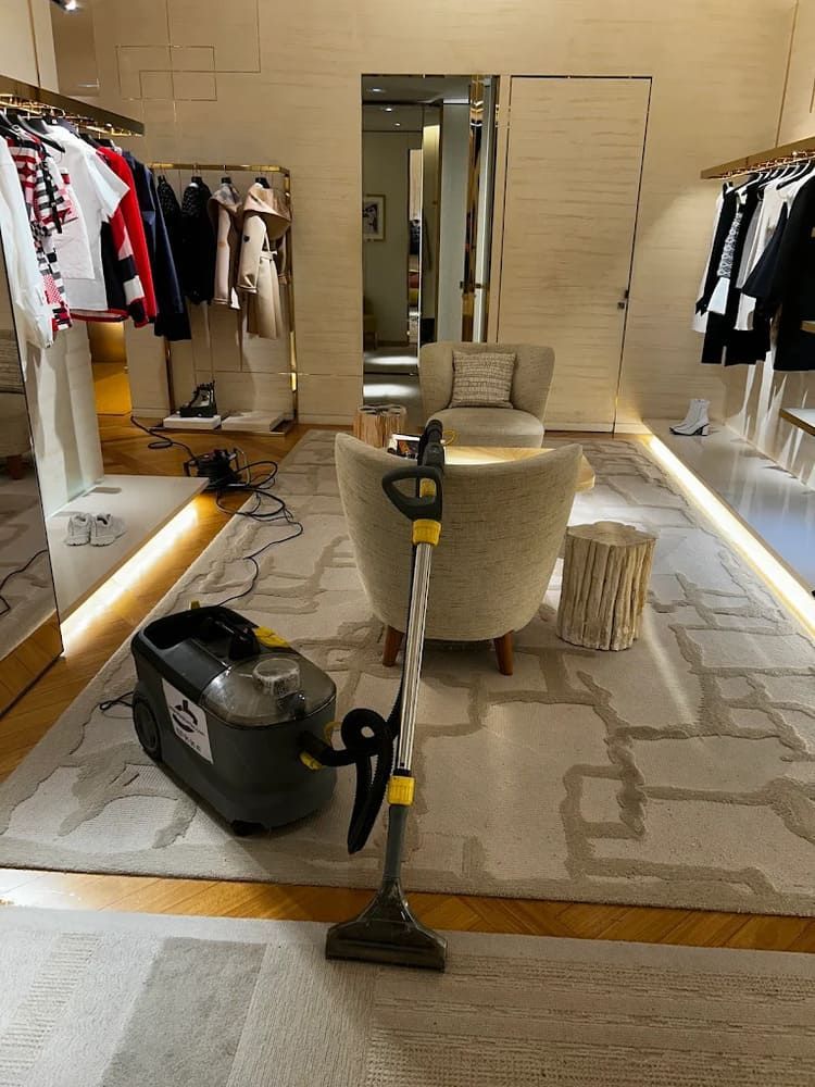 Una aspiradora limpia una alfombra beige en una tienda de ropa. La ropa está expuesta en percheros.