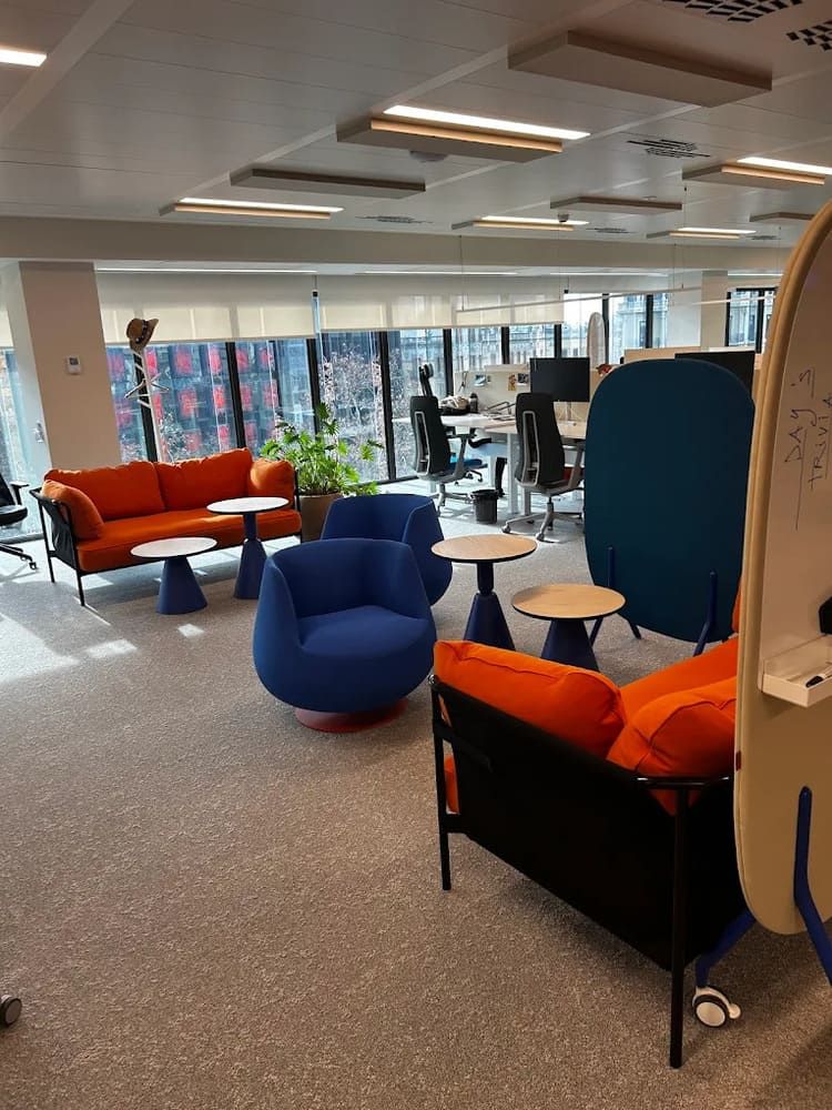 Zona de salón de oficina con muebles de color naranja y azul, mesas pequeñas y grandes ventanales.