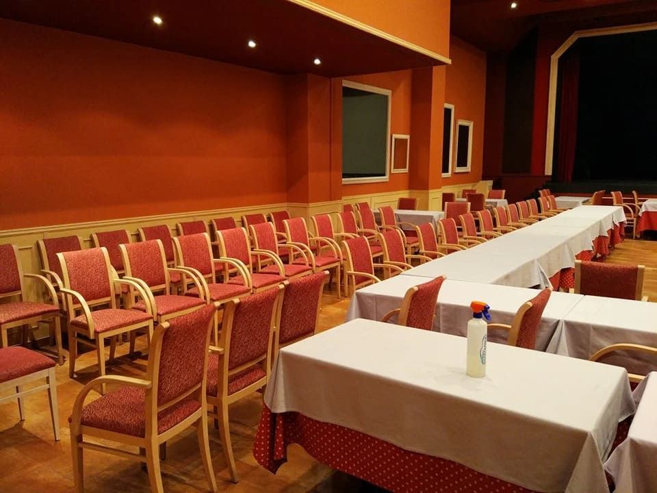 Filas de sillas rojas frente a largas mesas cubiertas con manteles blancos en una habitación con paredes naranjas.